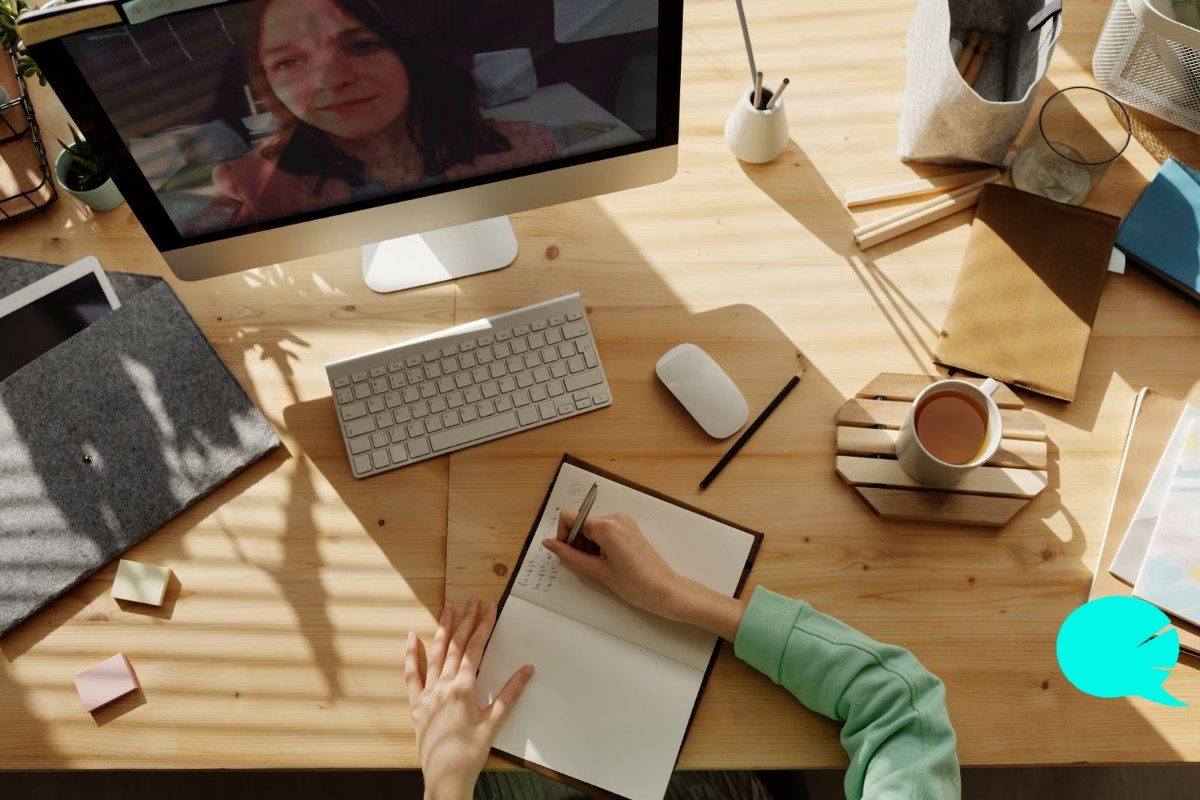 Mesa de trabajo con una videollamada de clase de inglés online en pantalla y una persona tomando apuntes